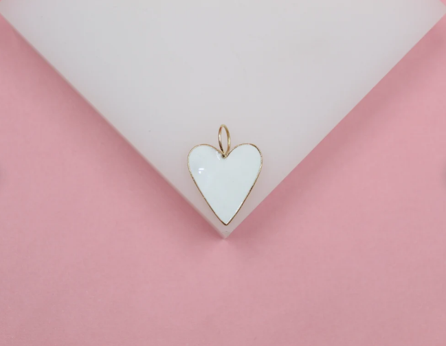 A gold-trimmed white enamel heart charm rests on a white display against a pink background.