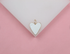 A gold-trimmed white enamel heart charm rests on a white display against a pink background.