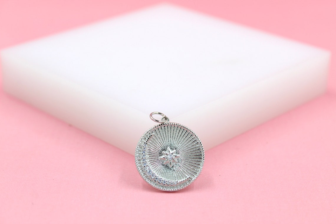 A round silver pendant featuring a crescent moon and star design rests on a white display block against a soft pink background.