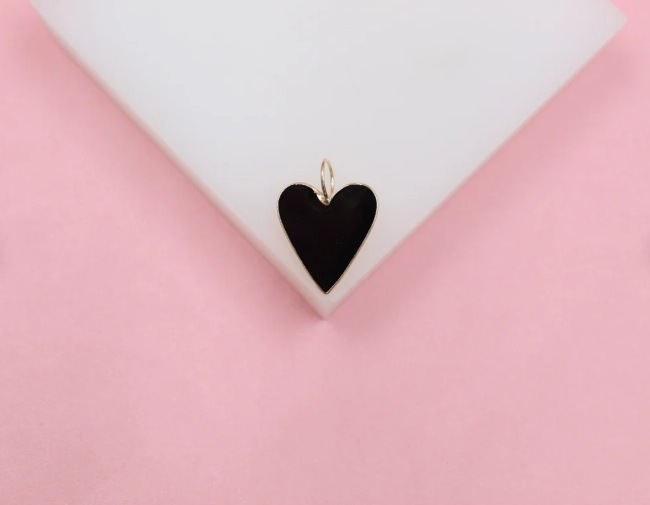 A gold-rimmed black enamel heart charm rests on a white display against a soft pink background.