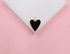 A gold-rimmed black enamel heart charm rests on a white display against a soft pink background.