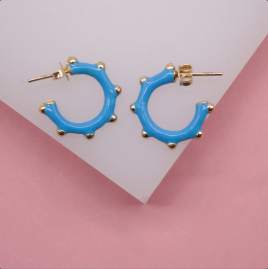 A pair of bright blue enamel hoop earrings with gold-plated stud backings rests on a white display against a pink background.