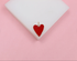 A glossy red enamel heart charm rests on a white display block against a soft pink background.