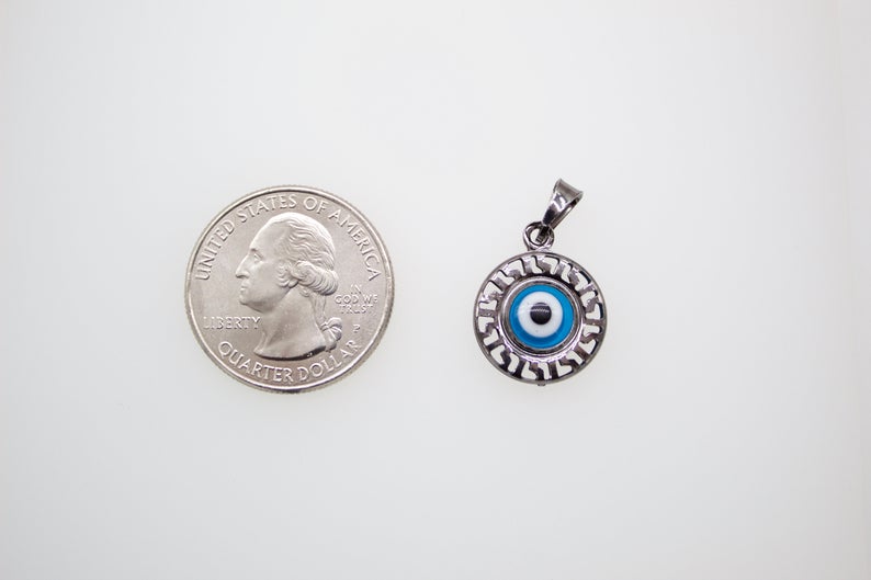 A silver evil eye pendant with Hebrew lettering rests next to a US quarter coin for scale on a white surface.