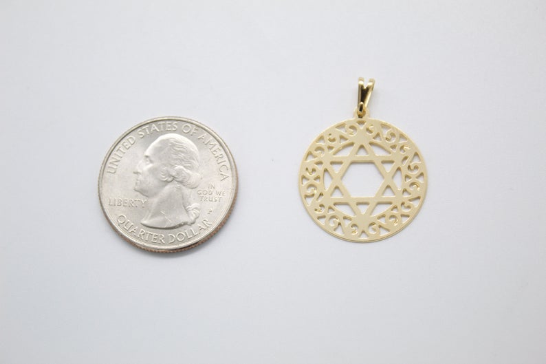 A round gold Star of David pendant rests beside a US quarter for size comparison on a light surface.