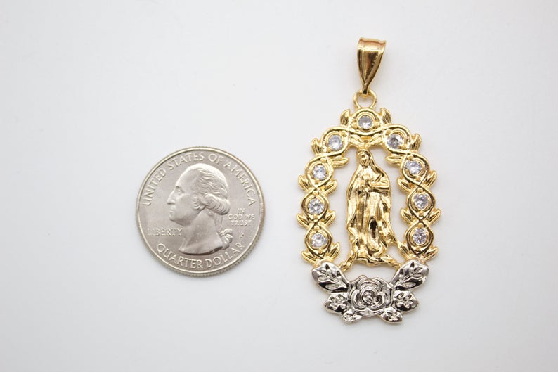 A gold Our Lady of Guadalupe pendant rests next to a US quarter coin for size comparison on a white surface.