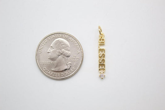 A gold 'Blessed' word charm with a diamond accent rests beside a US quarter for size comparison.