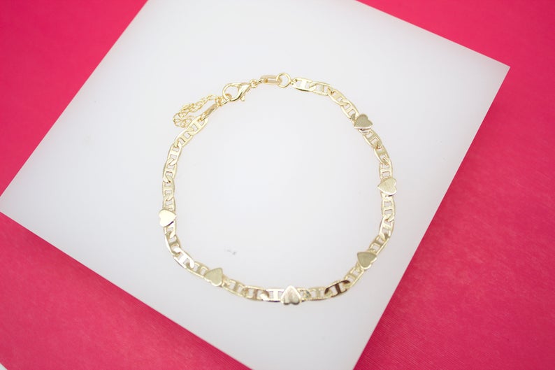 A delicate gold chain bracelet features alternating link patterns and small heart charms arranged in a circle on a white display surface against a vibrant pink background.
