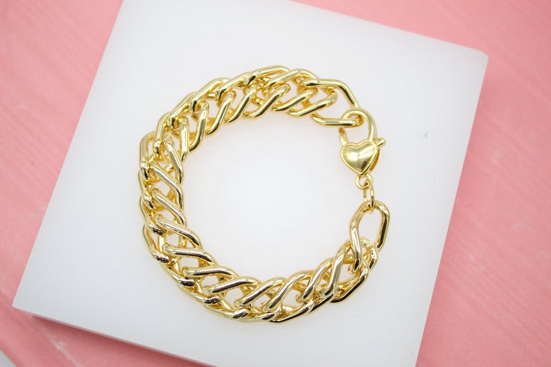 A thick gold curb chain bracelet with a heart-shaped clasp rests on a white display surface against a pink background.