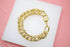 A thick gold curb chain bracelet with a heart-shaped clasp rests on a white display surface against a pink background.