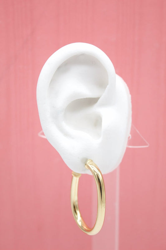 A thick gold hoop earring hangs from a white mannequin ear against a pink background.