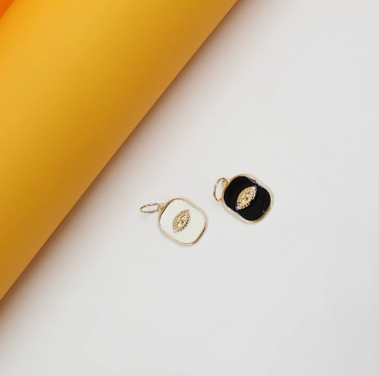 Two rectangular enamel charms featuring a gold evil eye motif rest on a white and yellow background.
