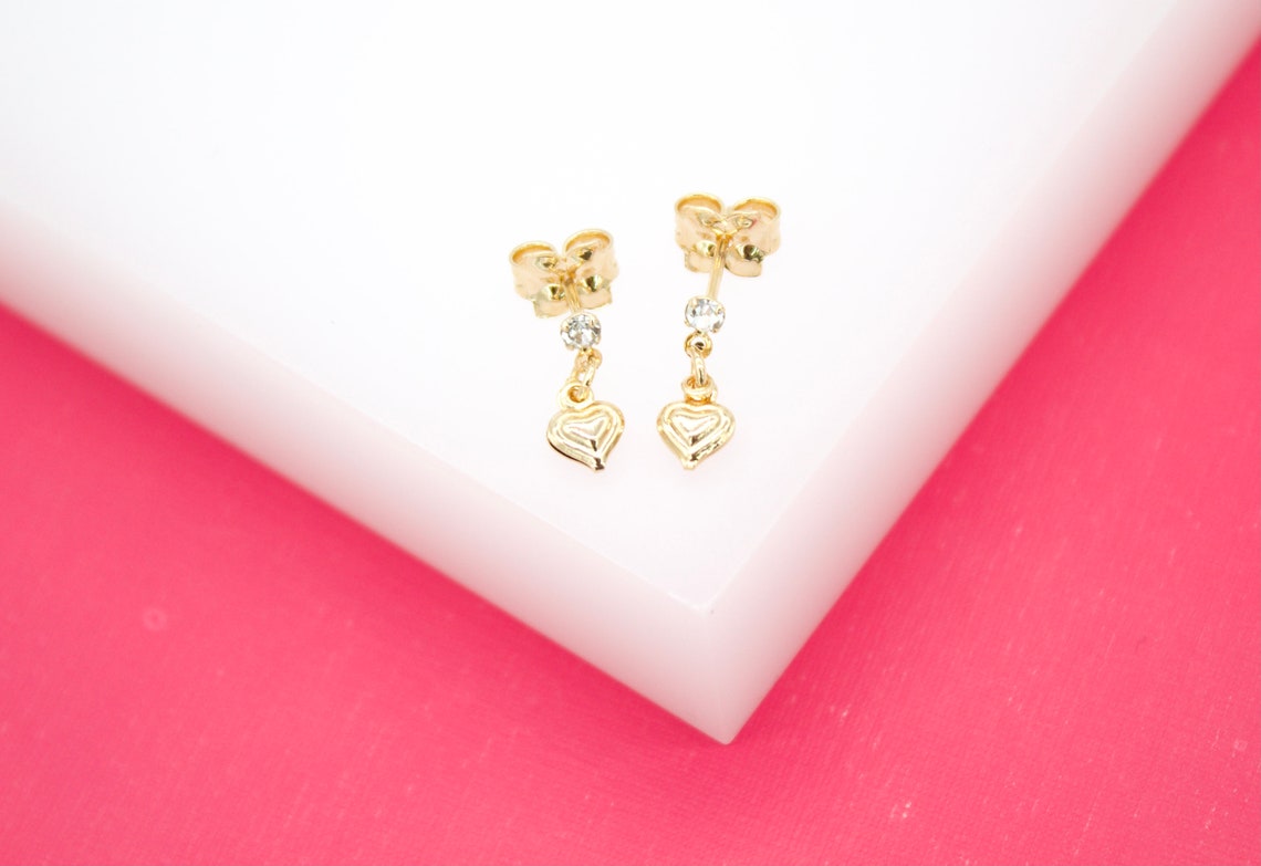 A pair of gold heart drop earrings rests on a white display block against a vibrant pink background.