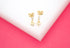 A pair of gold heart drop earrings rests on a white display block against a vibrant pink background.
