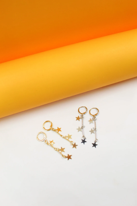 A pair of gold and a pair of silver star-dangle hoop earrings rest on a white surface against an orange backdrop.