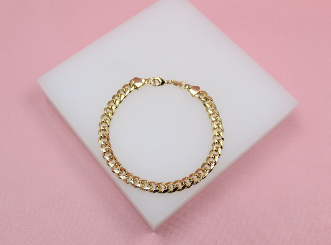A delicate gold cuban link bracelet rests on a white display block against a soft pink background.