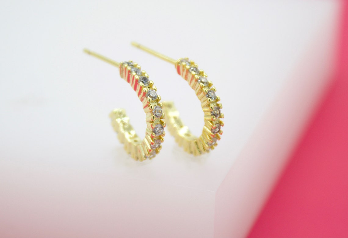 A pair of gold huggie hoop earrings embedded with small sparkling diamonds rest on a white surface against a bright pink background.