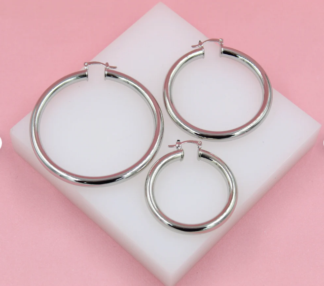 Three polished silver hoop earrings of varying sizes rest on a white display block against a soft pink background.