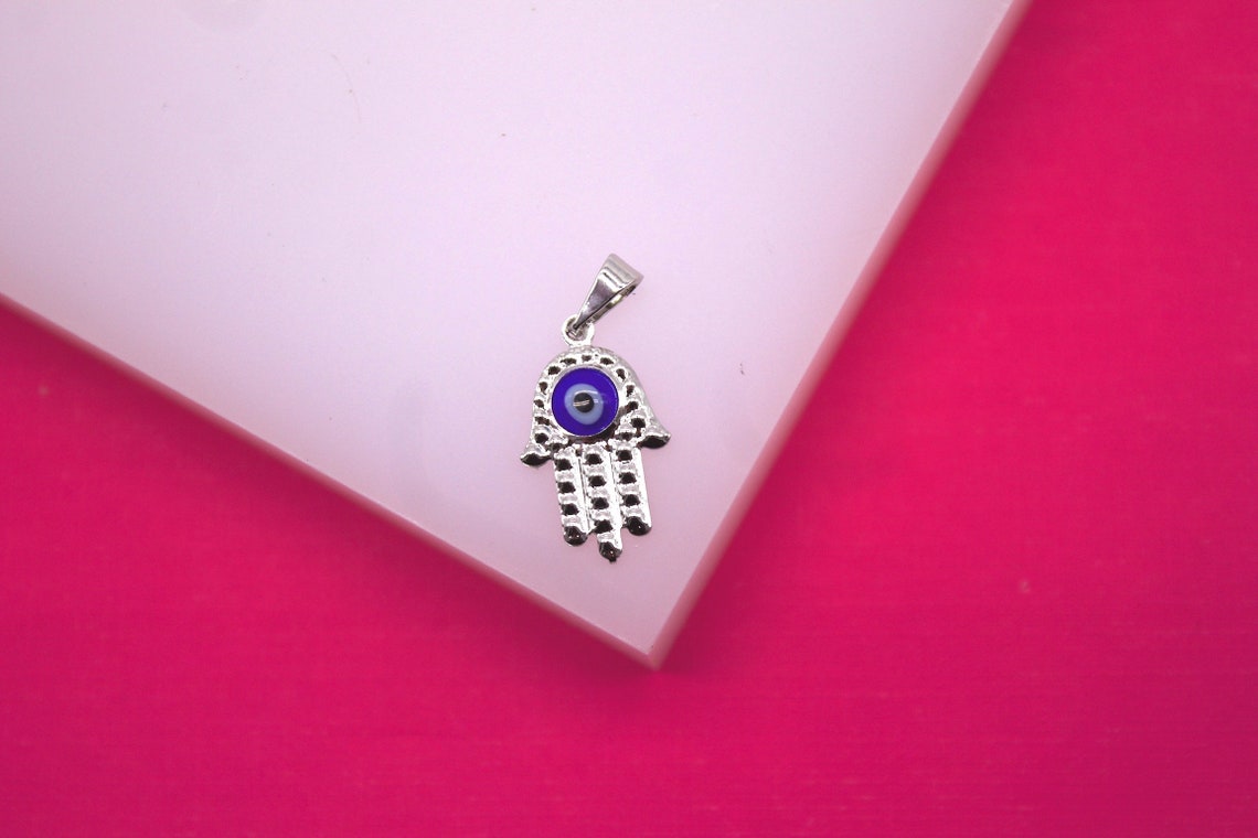 A silver Hamsa hand pendant featuring a blue evil eye rests on a white display against a vibrant pink background.