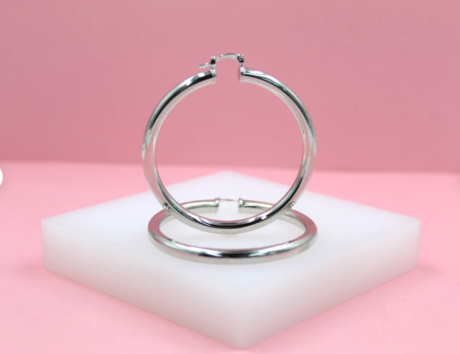 A pair of polished silver hoop earrings rests on a white display block against a soft pink background.