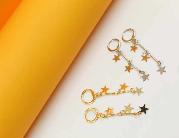 Gold and silver star-charm hoop earrings rest against a bright orange and white backdrop.