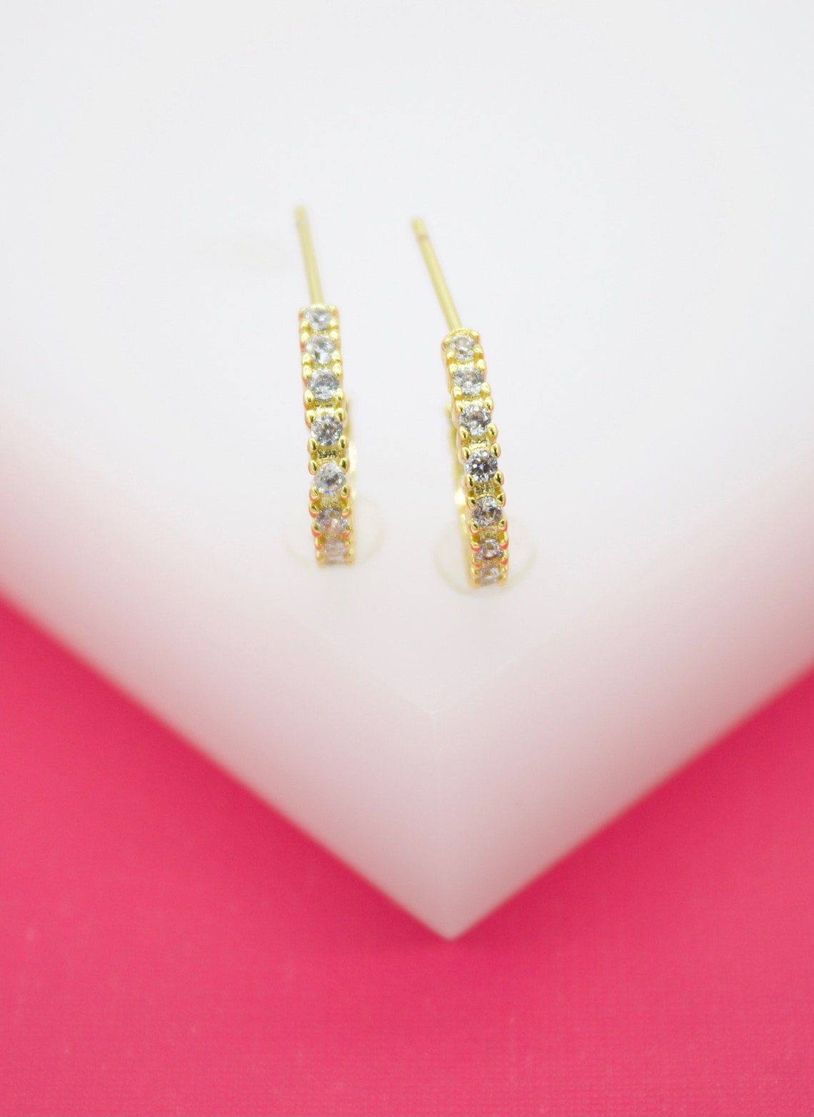 A pair of gold huggie hoop earrings embedded with sparkling cubic zirconia stones rests upright on a white display block against a vibrant pink background.