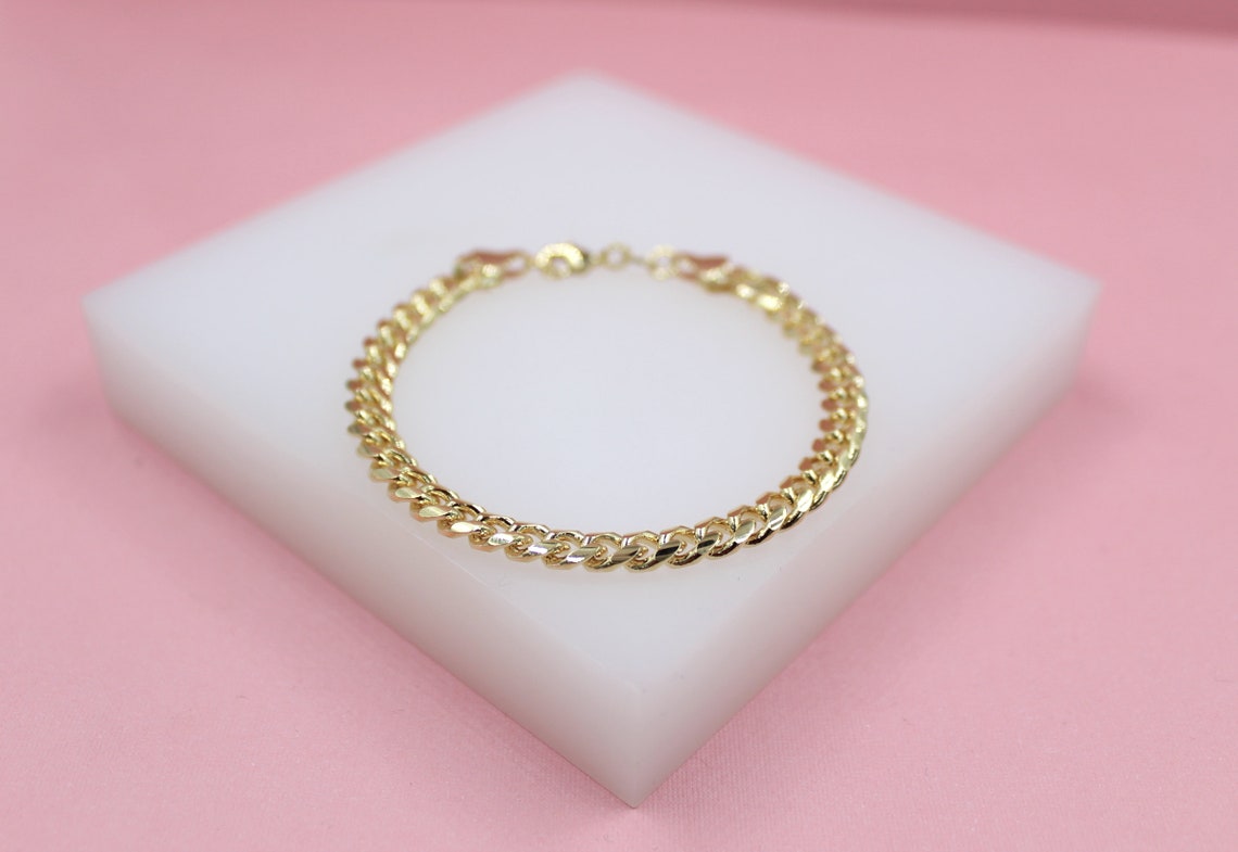 A shiny gold cuban link bracelet rests on a white display block against a soft pink background.