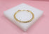 A shiny gold cuban link bracelet rests on a white display block against a soft pink background.