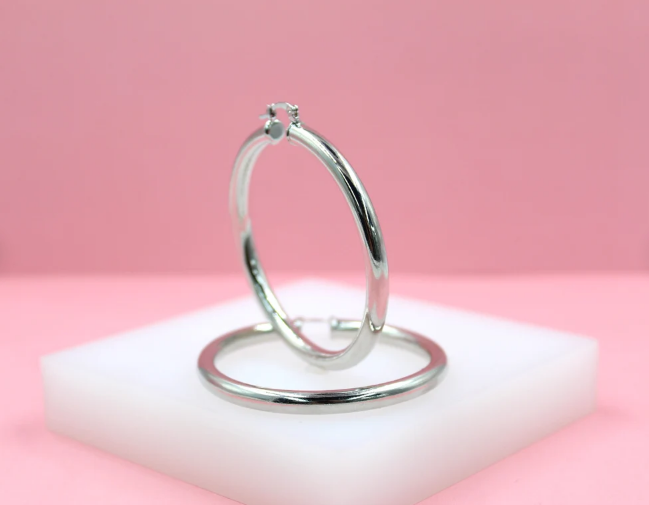 A pair of polished silver hoop earrings rests on a small white display block against a soft pink background.