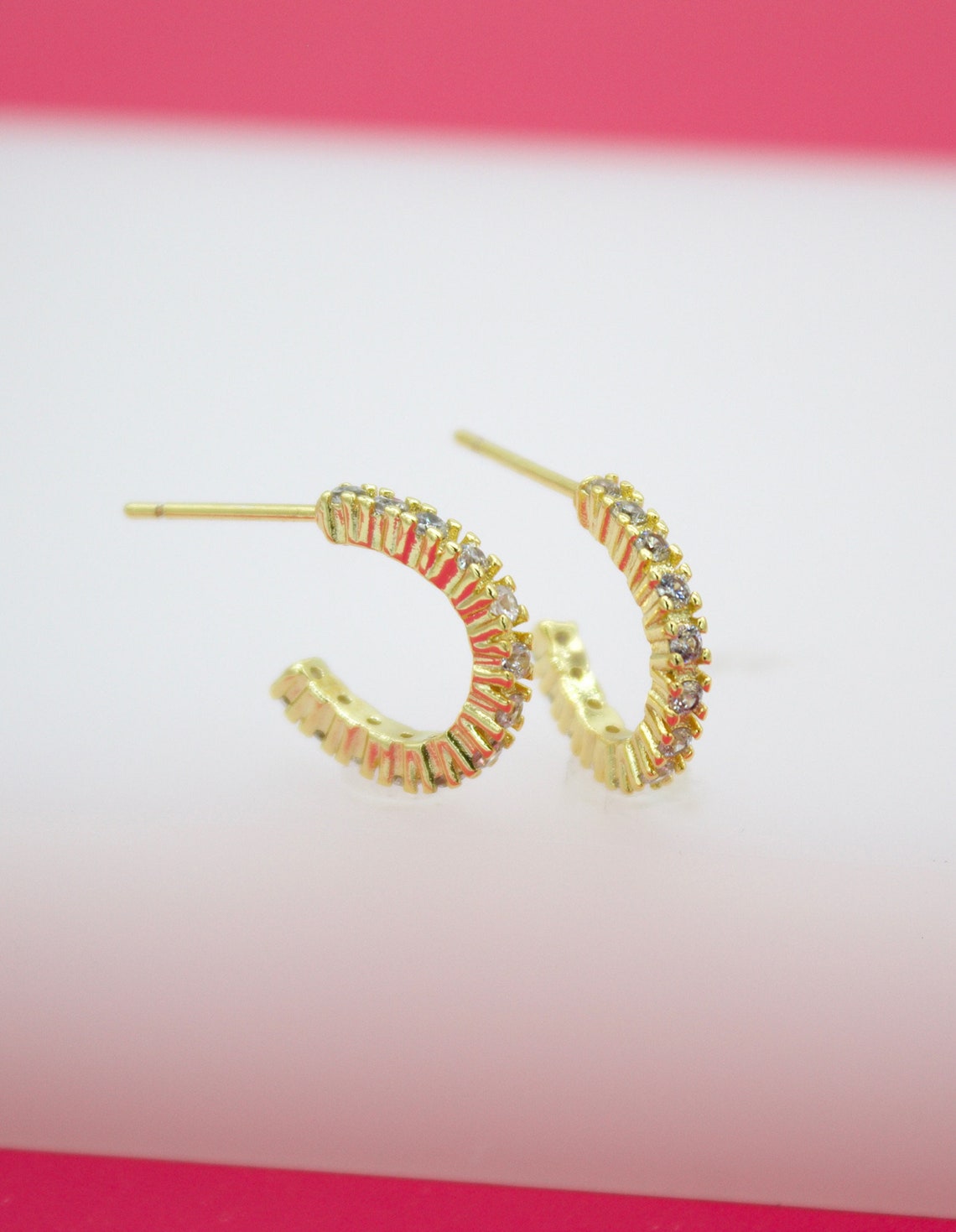 A pair of gold huggie hoop earrings embedded with sparkling cz stones rests on a white display surface against a pink background.