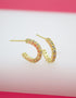 A pair of gold huggie hoop earrings embedded with sparkling cz stones rests on a white display surface against a pink background.