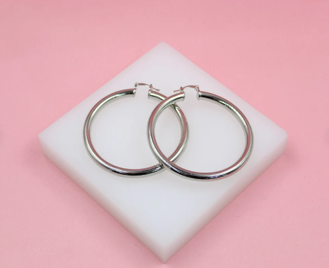 A pair of glossy silver hoop earrings rests on a white display block against a solid pink backdrop.