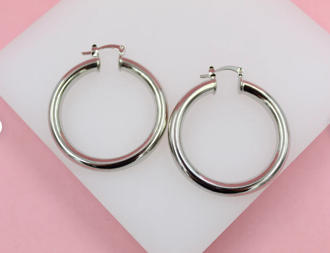 A pair of polished silver hoop earrings rests on a white display block against a soft pink background.