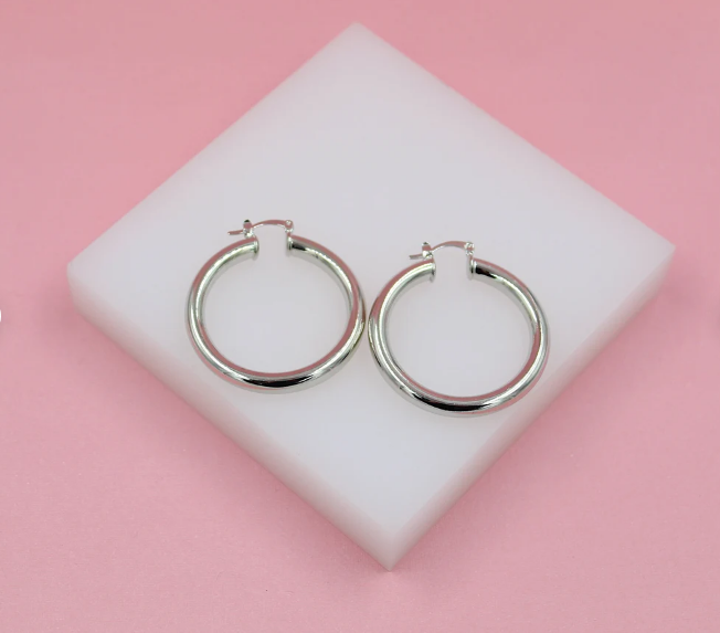 A pair of polished silver hoop earrings rests on a white block against a soft pink background.