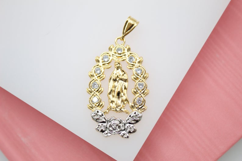 A gold Our Lady of Guadalupe pendant rests on a white and pink surface, featuring a sparkling halo of cubic zirconia stones and a silver rose detail at the base.