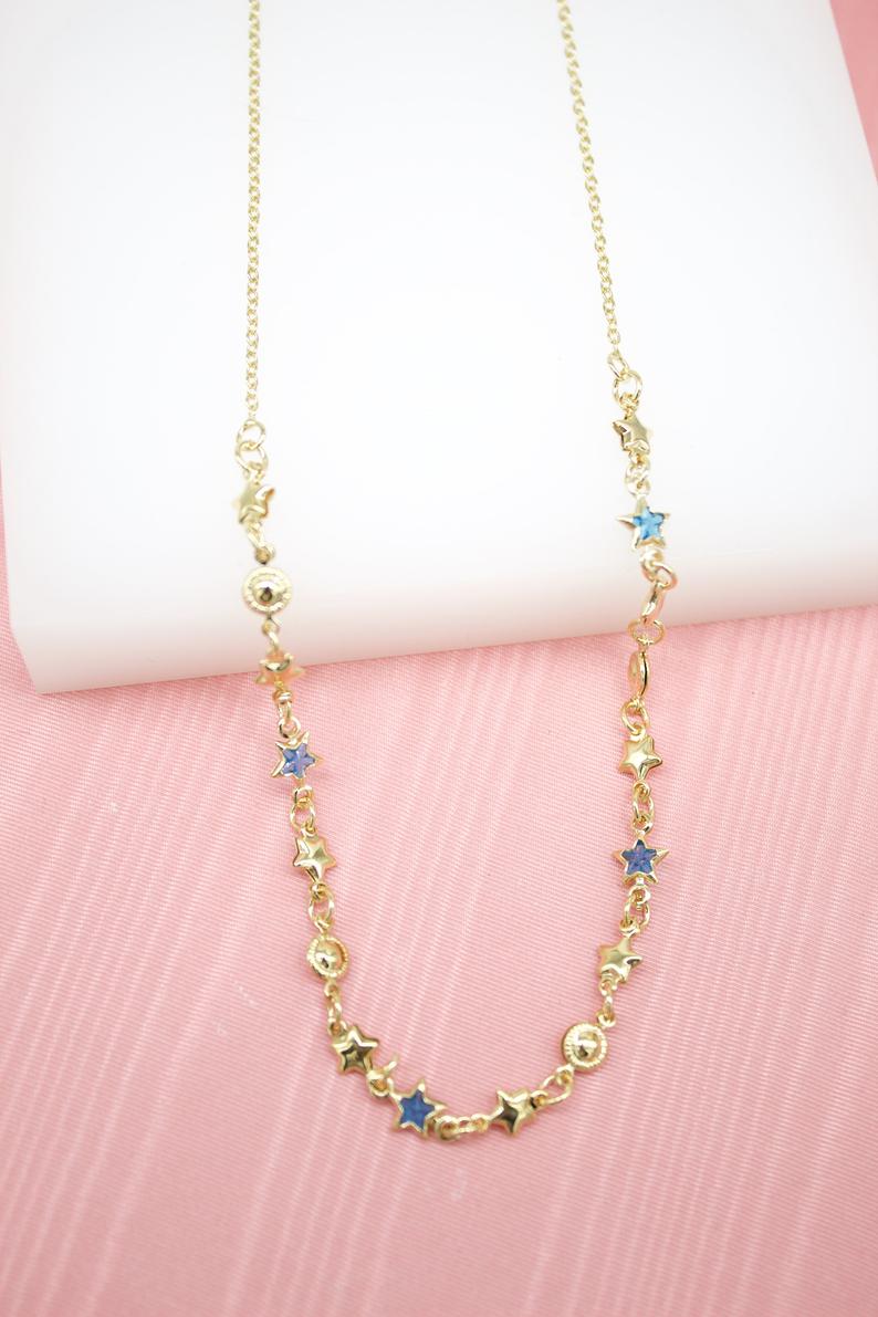 A delicate gold star pendant necklace rests against a soft pink backdrop, featuring scattered blue enamel star charms along its chain.