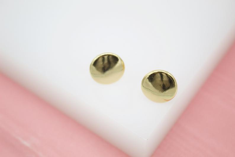 A pair of shiny gold-plated round stud earrings rests on a white display against a soft pink background.