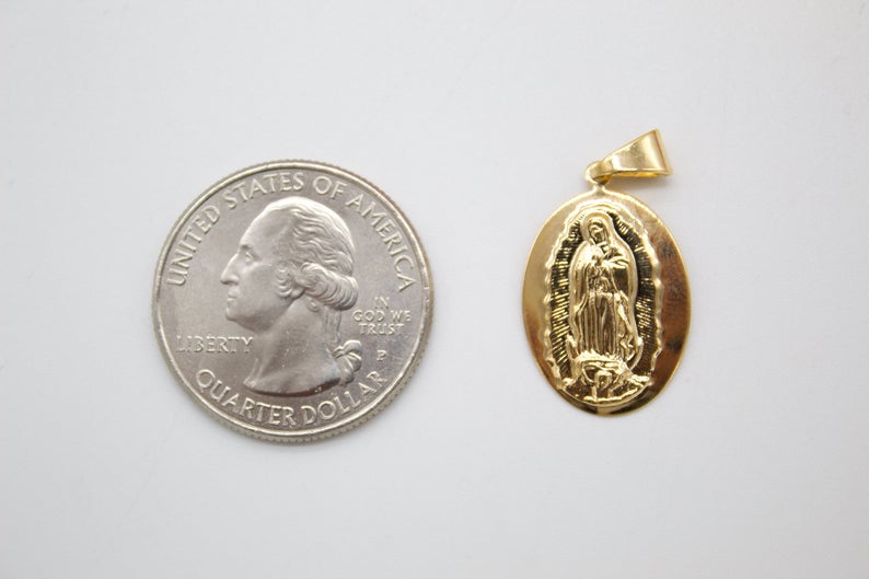 A gold oval Our Lady of Guadalupe pendant rests next to a US quarter coin for a size comparison on a plain surface.