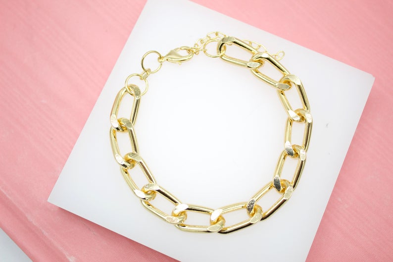 A thick gold chunky link bracelet rests on a white display surface against a pink background.