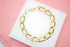 A thick gold chunky link bracelet rests on a white display surface against a pink background.