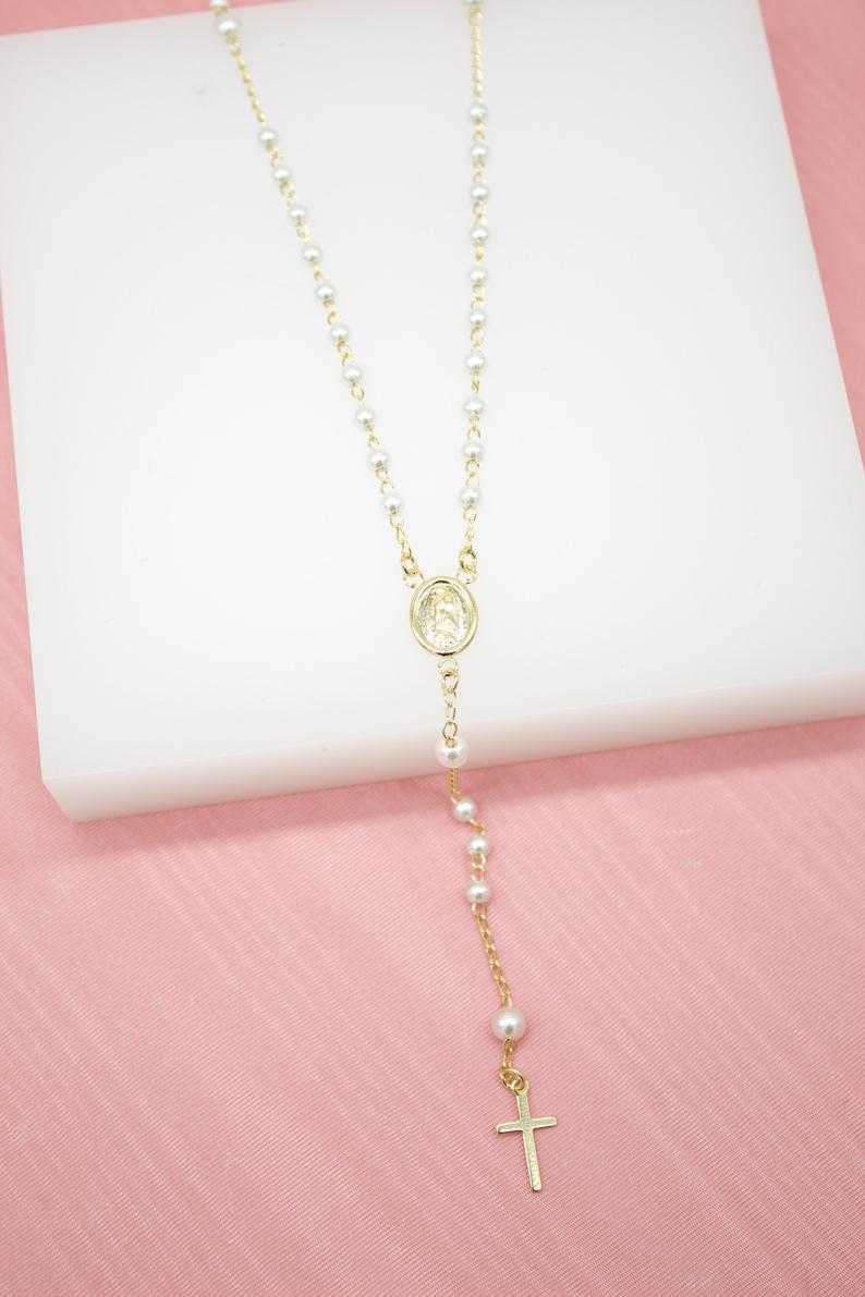 A delicate gold rosary necklace featuring white pearls and a matching crucifix rests on a white display surface against a pink background.