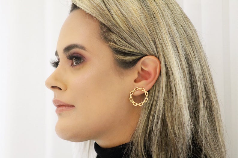 A blonde woman wearing a stylish gold chain-link hoop earring gazes thoughtfully in profile.