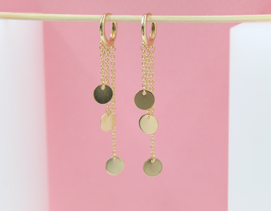 A pair of delicate gold chain earrings with hanging coin charms hangs from a thin wooden stick against a pink and white background.