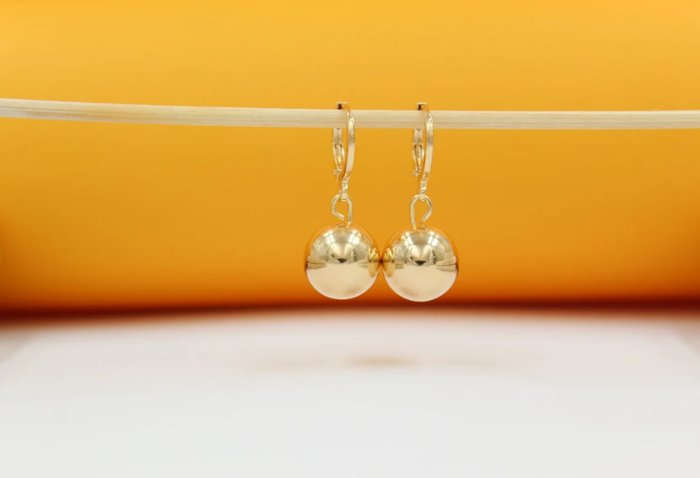 A pair of shiny gold ball drop earrings hangs delicately from a thin stick against a warm orange background.