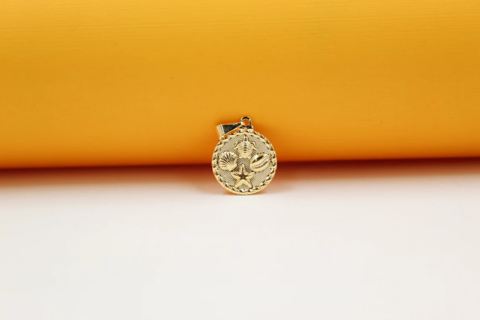 A gold pendant featuring a circular medallion with embossed sea life motifs, including a starfish, turtle, and seashells, rests against a vibrant yellow and white background.