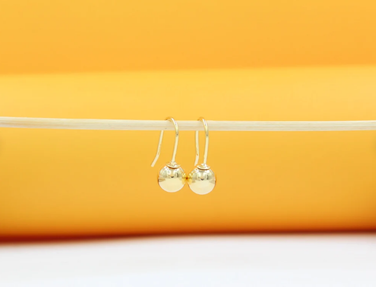 A pair of shiny gold ball drop earrings hangs delicately from a thin stick against a solid yellow background.