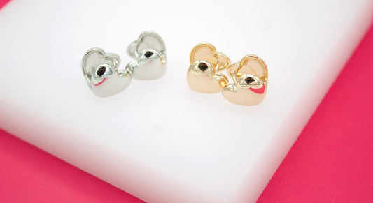 A pair of silver heart-shaped huggie earrings sits next to a matching pair of gold heart-shaped huggie earrings on a white and pink display surface.