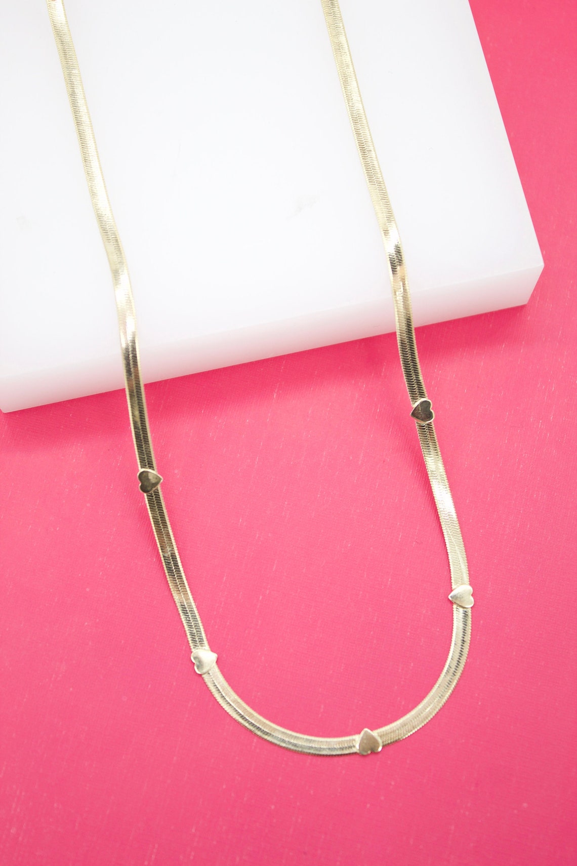 A flat gold herringbone chain necklace adorned with small heart charms rests against a white display block and vibrant pink background.