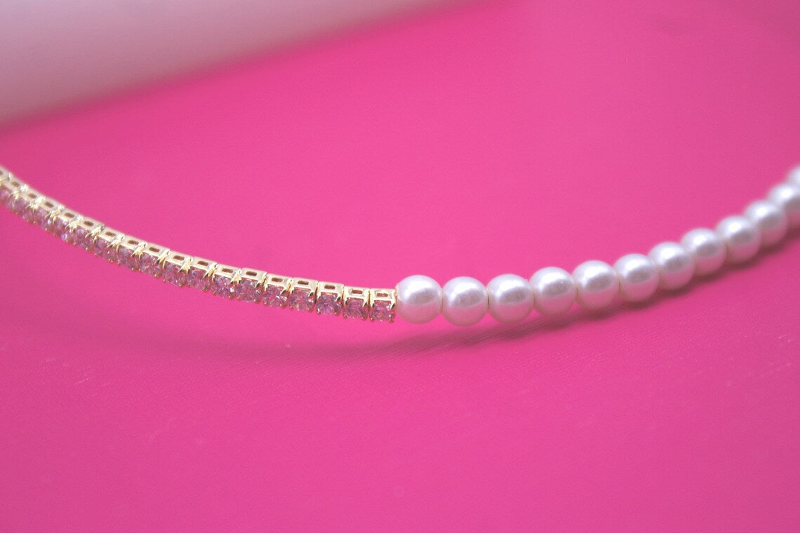 A stylish necklace combines a row of sparkling pink crystal gemstones on one side with classic white pearls on the other against a vibrant pink background.