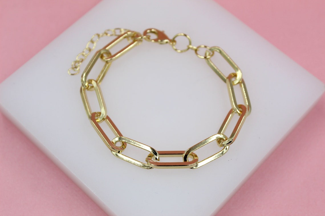 A shiny gold paperclip chain bracelet rests on a white display block against a soft pink background.